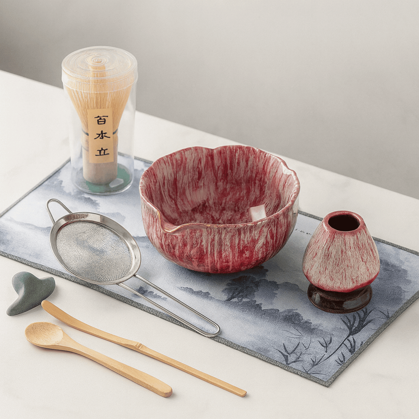 Red ceramic bowl with matching small pitcher on a decorative mat with tea-making tools.