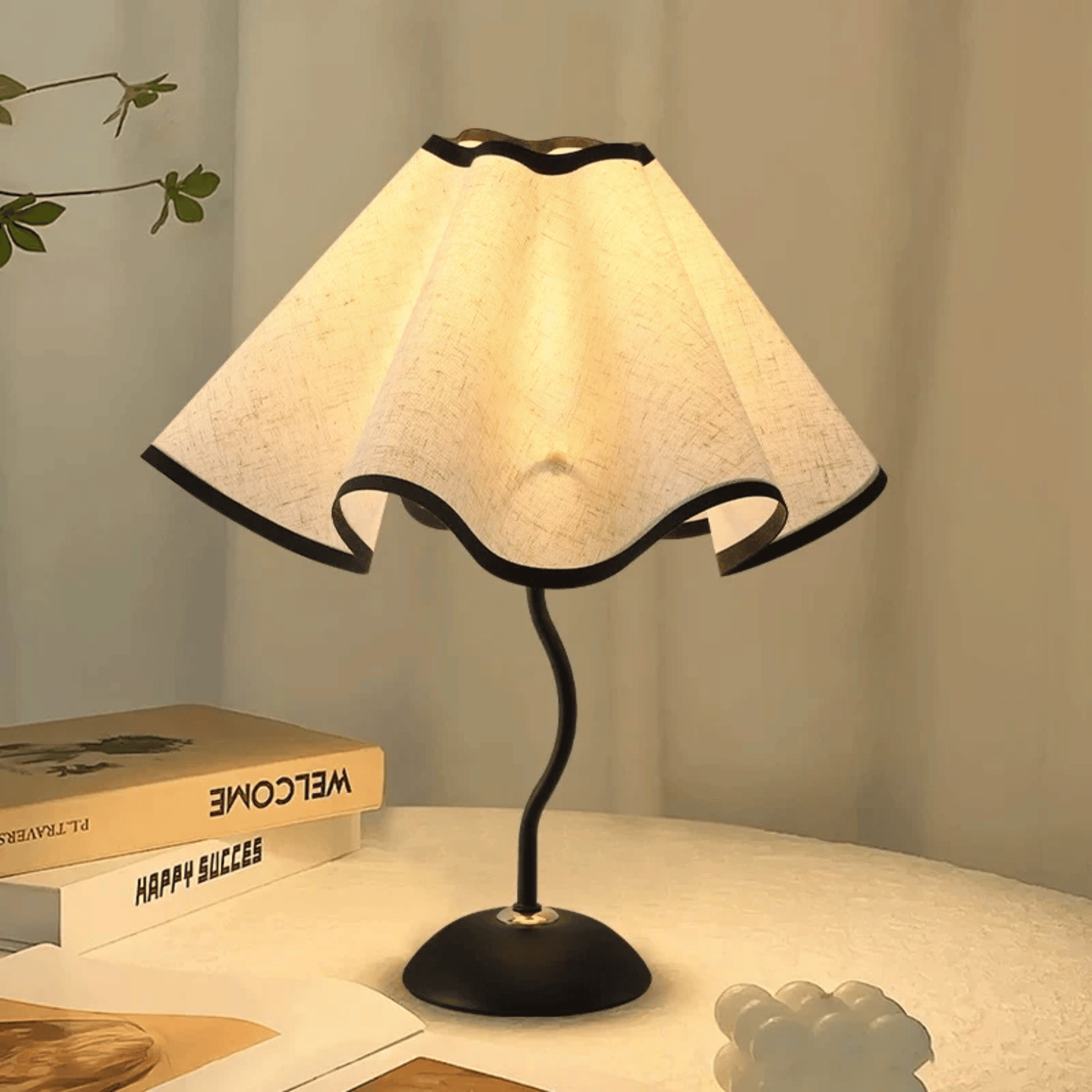 Table lamp with a white shade on a surface with books and a plant in the background
