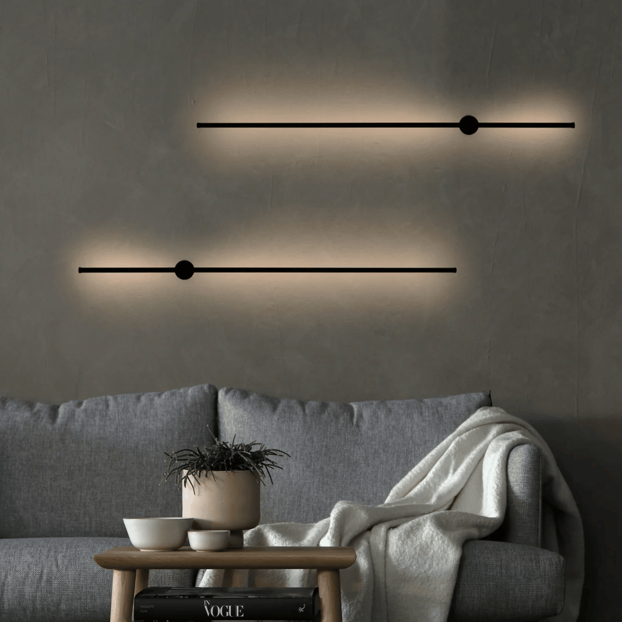 Modern wall lights on a gray wall above a gray sofa with a small table and plant.
