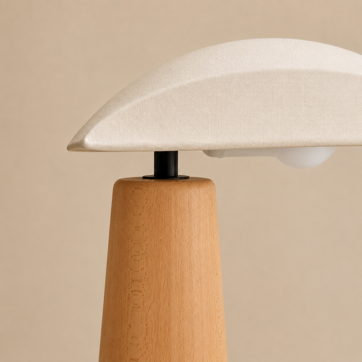 A close-up of a table lamp with a light beige fabric dome shade, a natural wood tapered base, and a black metal connector, set against a solid beige background.