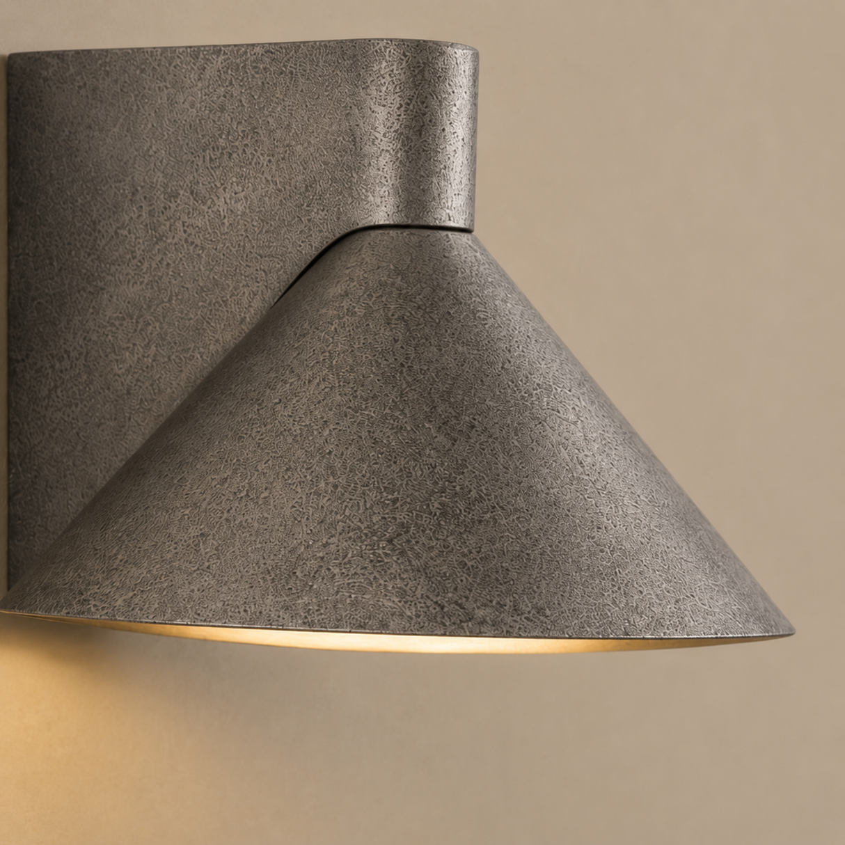 A modern wall-mounted sconce with a textured gray metal shade and a warm light glowing from within, shown against a beige wall.