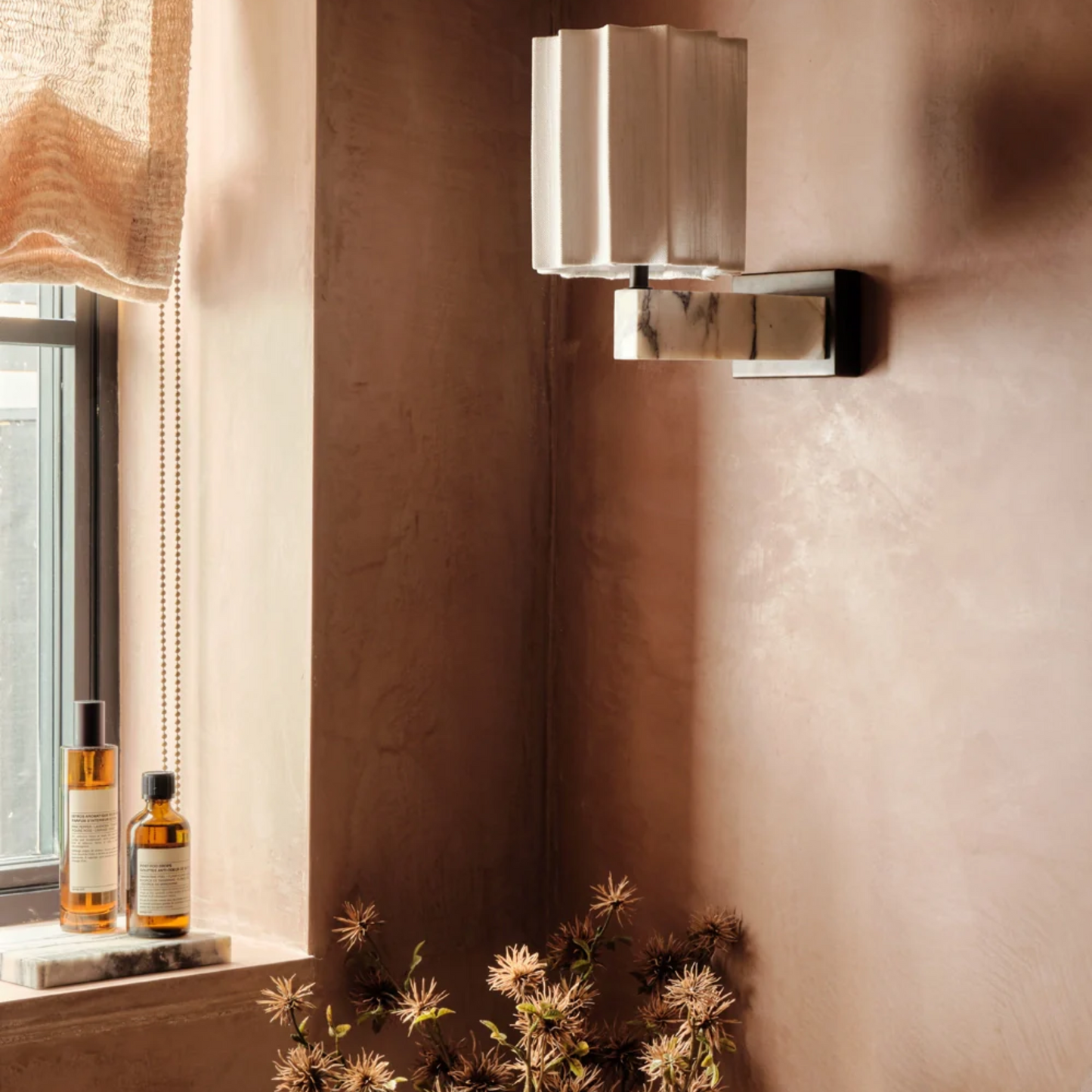 Modern wall sconce on a textured wall with bottles and plants below a window.