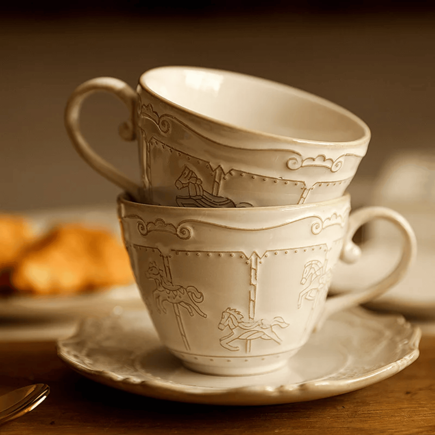 Two stacked ceramic cups with decorative patterns on a saucer, with pastries in the background.