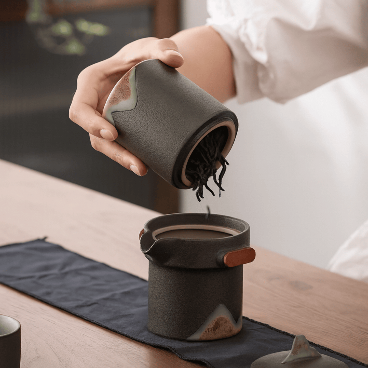 Person pouring from a ceramic container into another on a wooden table.