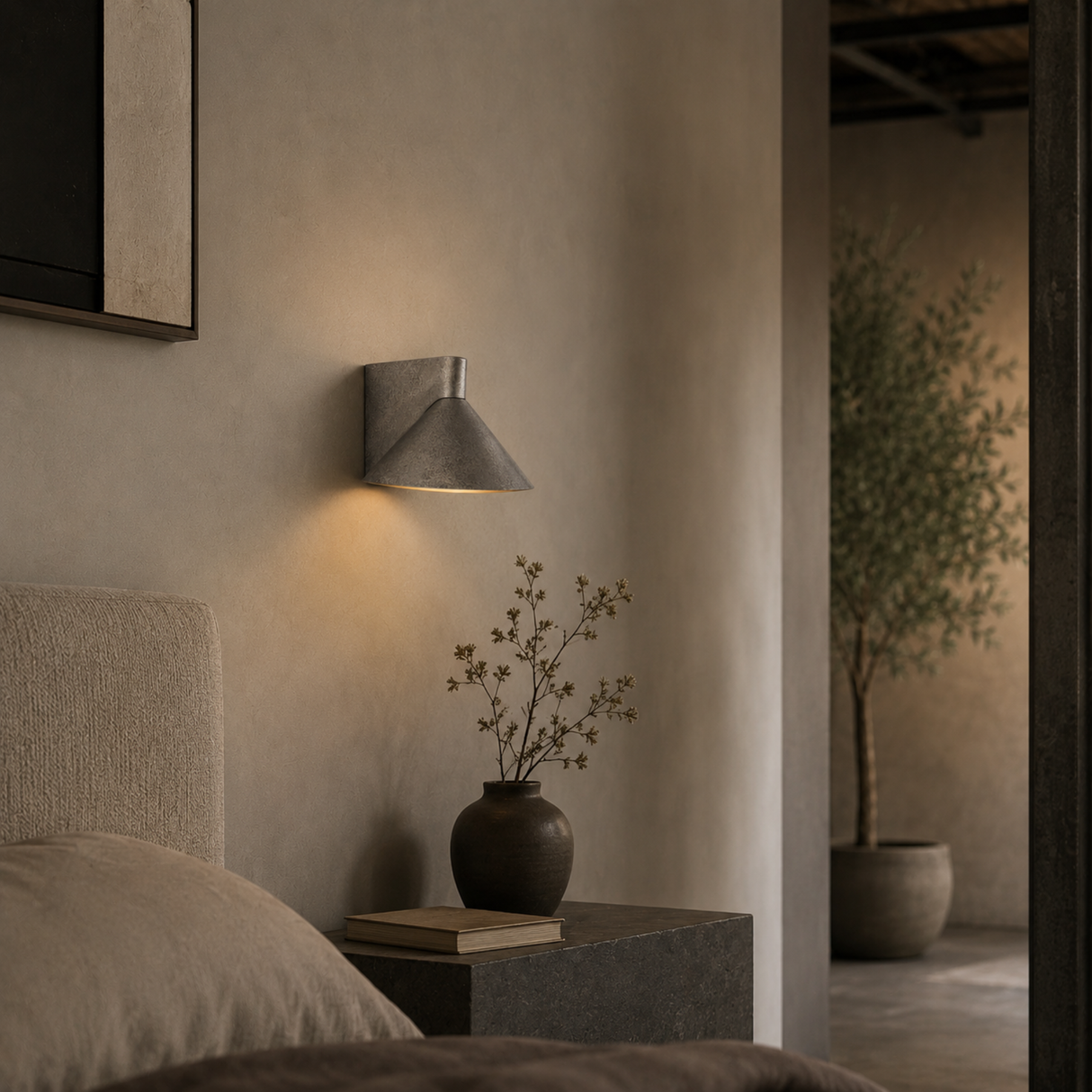 A modern wall-mounted sconce with a conical metal shade, casting a warm light on a textured beige wall above a bedside table with a vase and books.