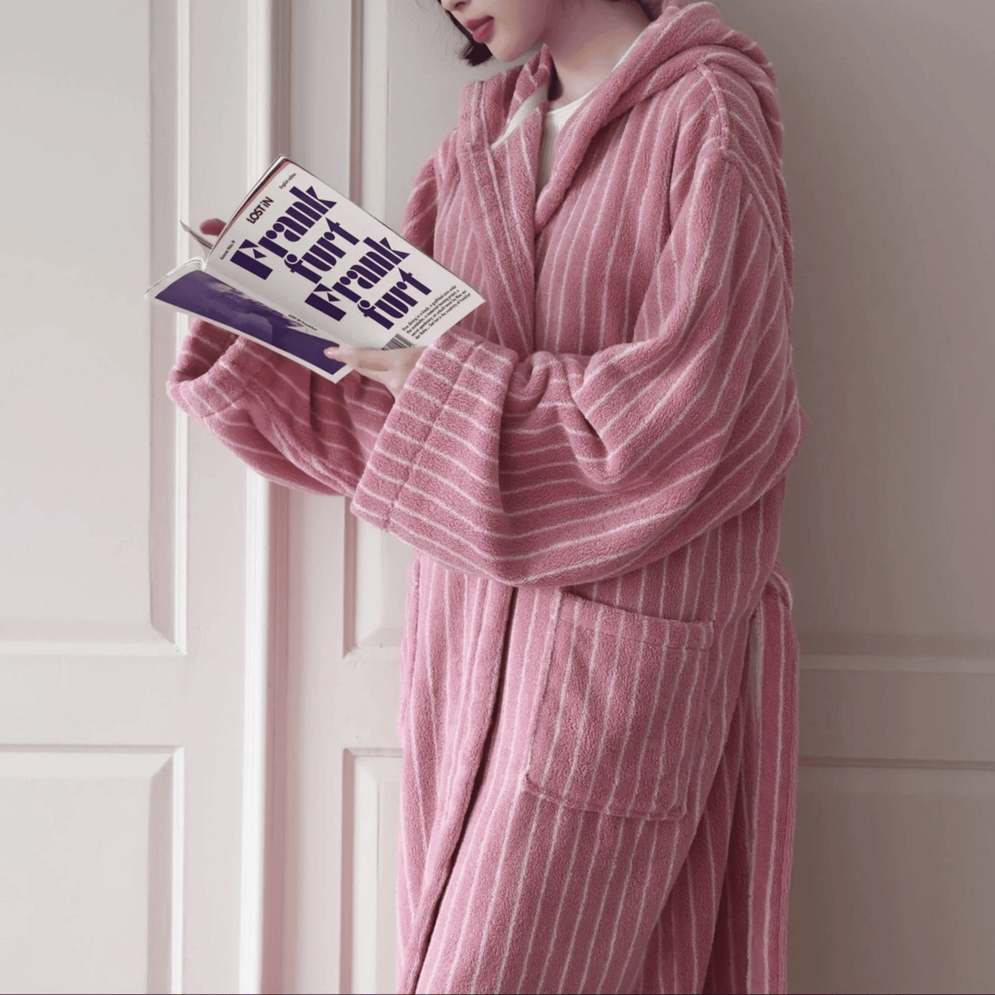 Person wearing a pink striped robe holding a book in a room with white paneled walls.