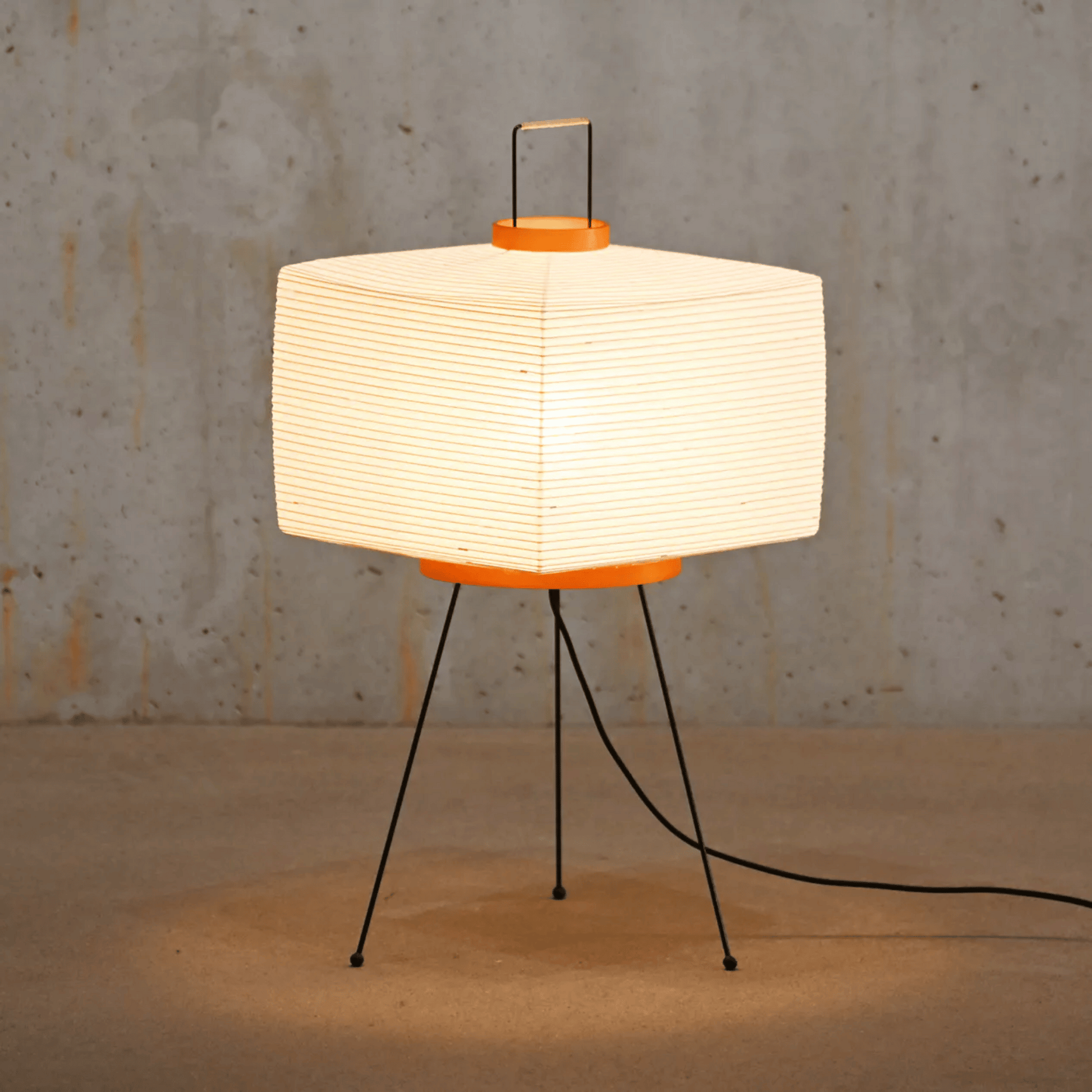 Table lamp with a white paper shade and orange base on a concrete floor.