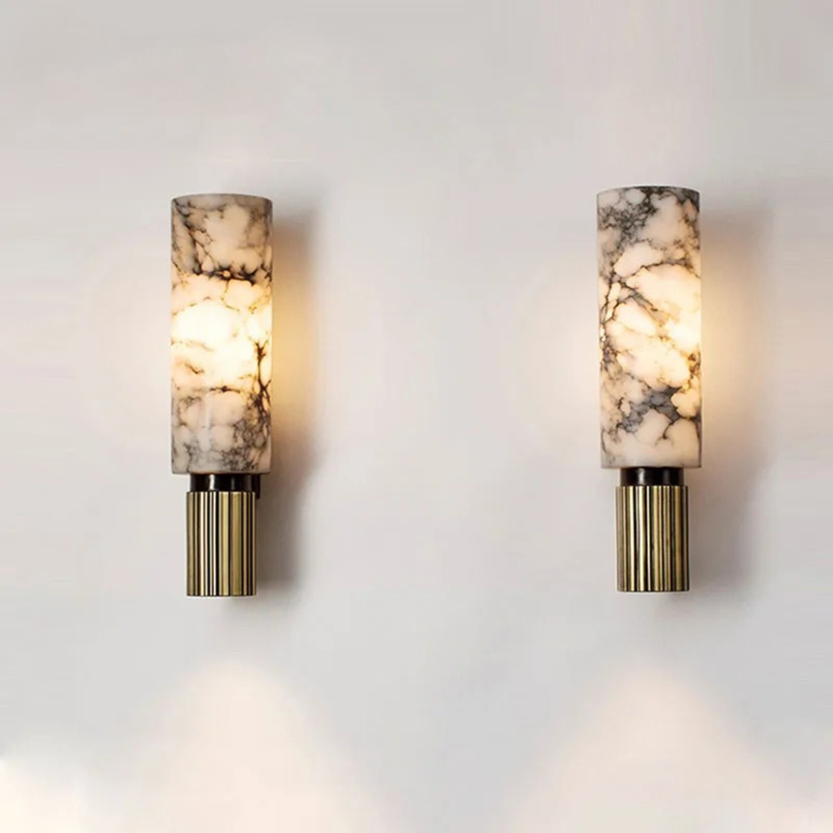 Two modern wall sconces with cylindrical, marble-patterned glass shades and gold ribbed bases are mounted on a plain white wall, emitting a warm, soft light.