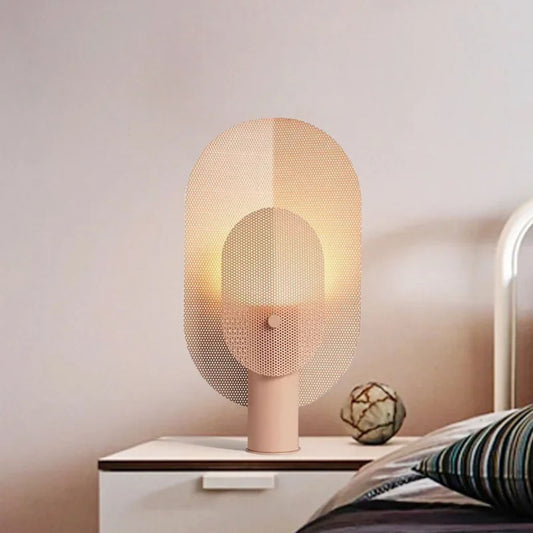 A modern table lamp with a soft pink base and a woven mesh shade, casting a warm glow on a bedside table.