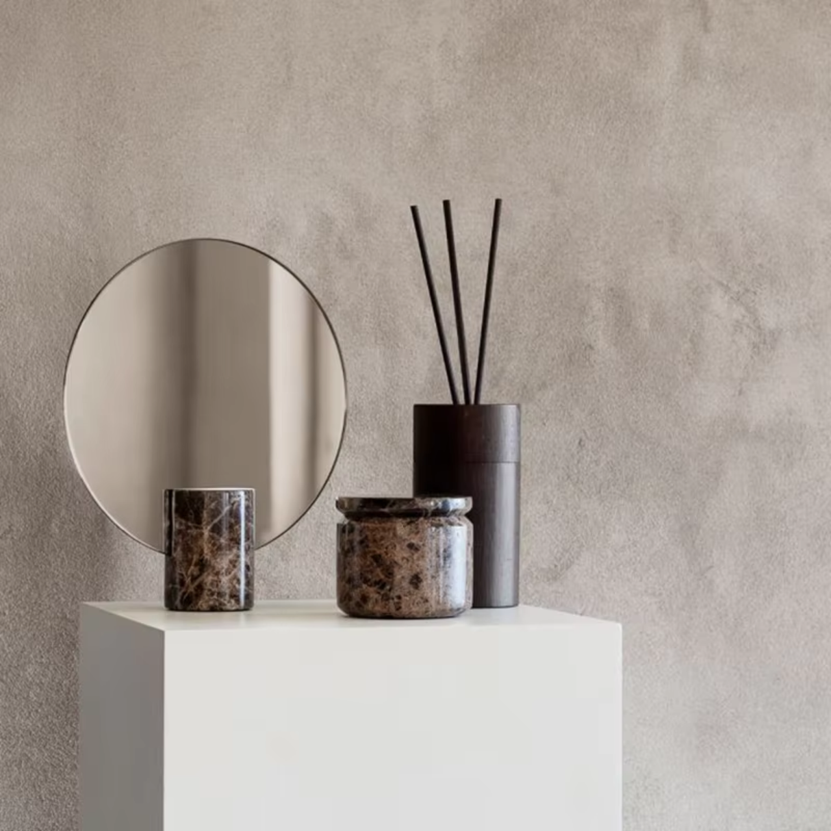 Round mirror, candles, and diffuser on a white pedestal against a beige wall.