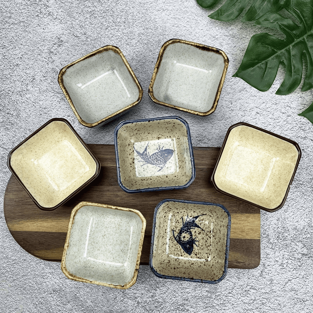 Set of square ceramic dishes with fish design on a wooden board, on a textured surface with leaves.