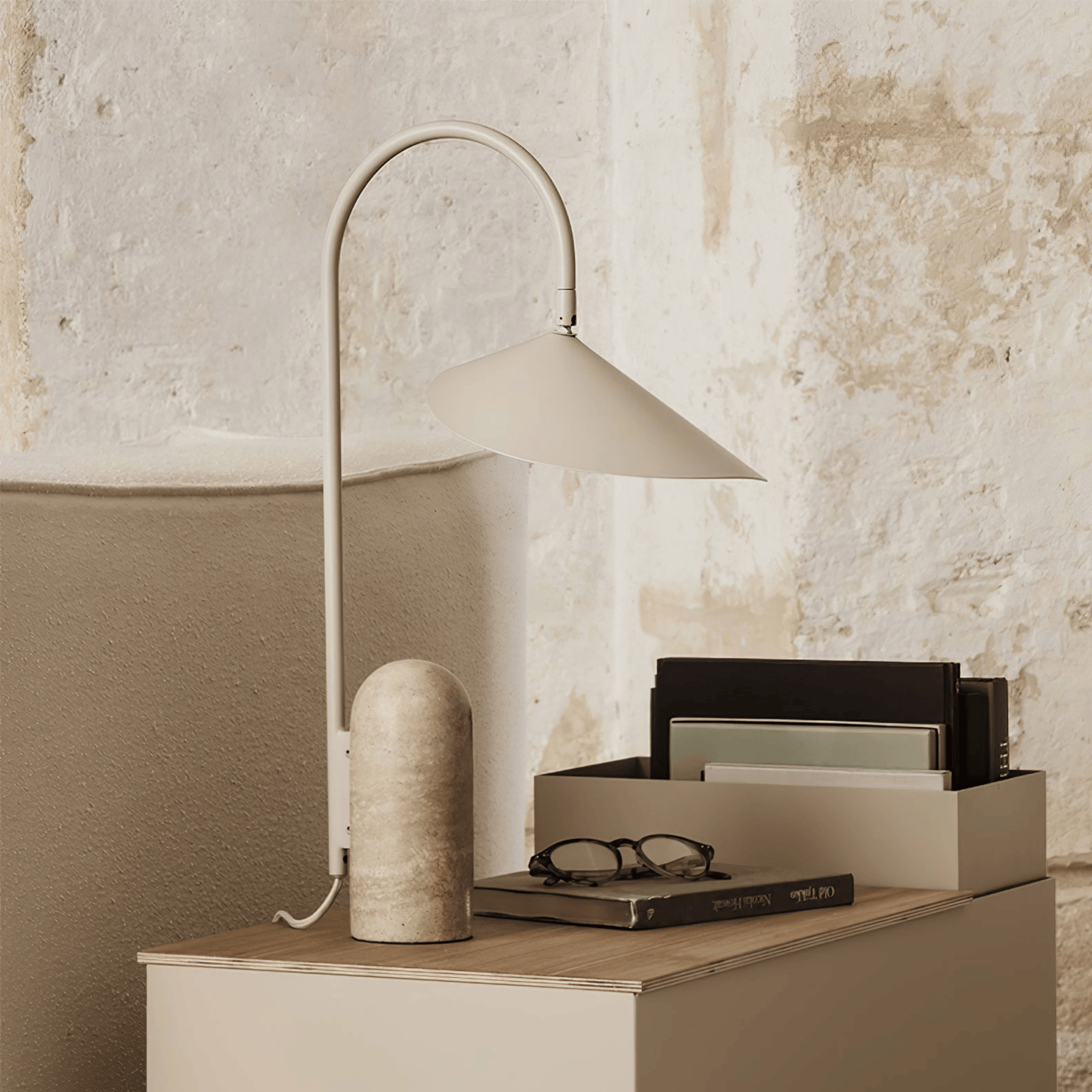 Beige table lamp on a wooden surface with books and glasses against a textured wall.