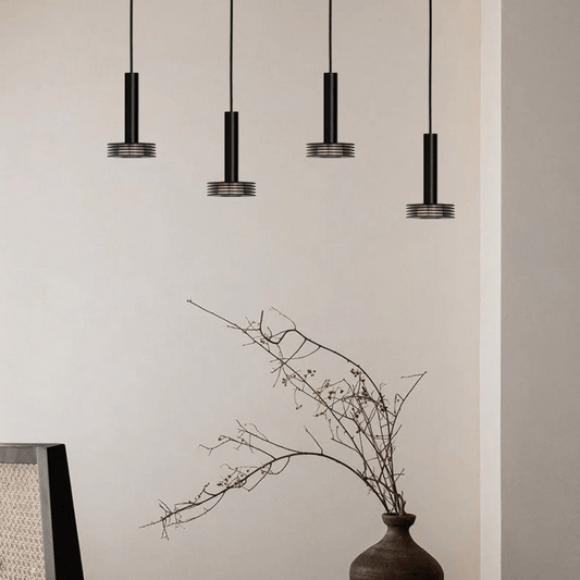 Four black pendant lights hanging against a beige wall with a vase of branches on a table.