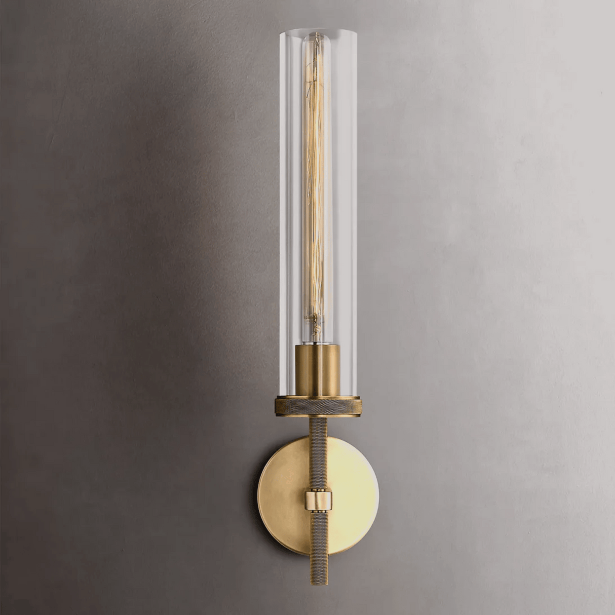 Modern wall sconce with clear glass shade and brass finish on a gray wall.