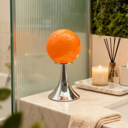 An orange glass globe table lamp on a chrome base displayed on a light-colored tabletop near a window.