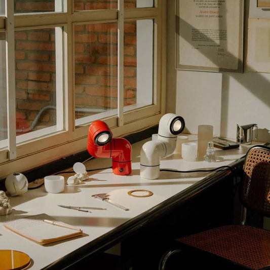 A red and white desk lamp designed as elbow pipes, placed on a table with a mix of items including a pen, notebook, and cups, near a window.