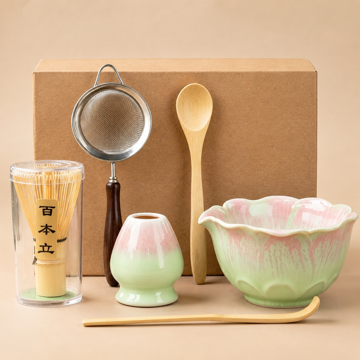 Ceramic tea set with a pink and green flower-shaped bowl, wooden spoon, metal sieve, bamboo whisk in a glass holder, and a small vase, arranged in front of a brown box on a beige background.