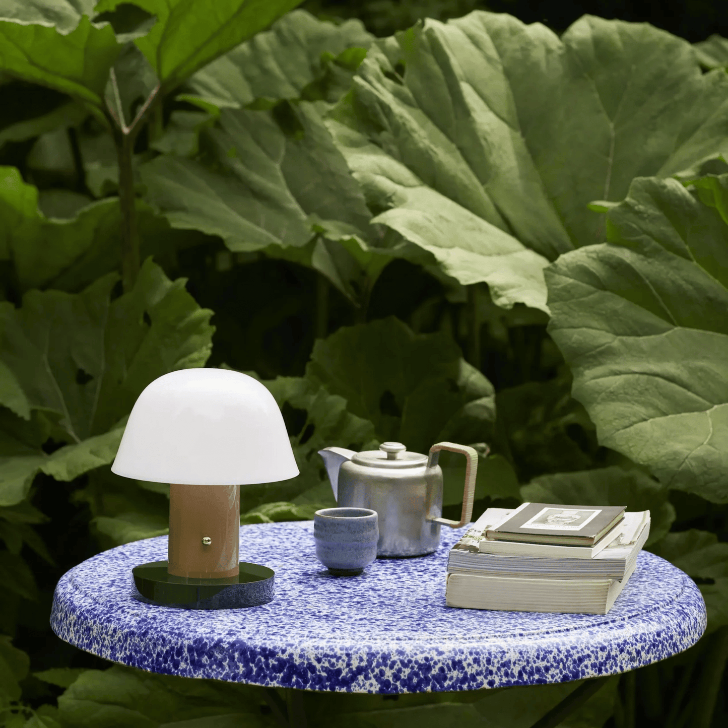 Table with a lamp, teapot, and books against a green leafy background