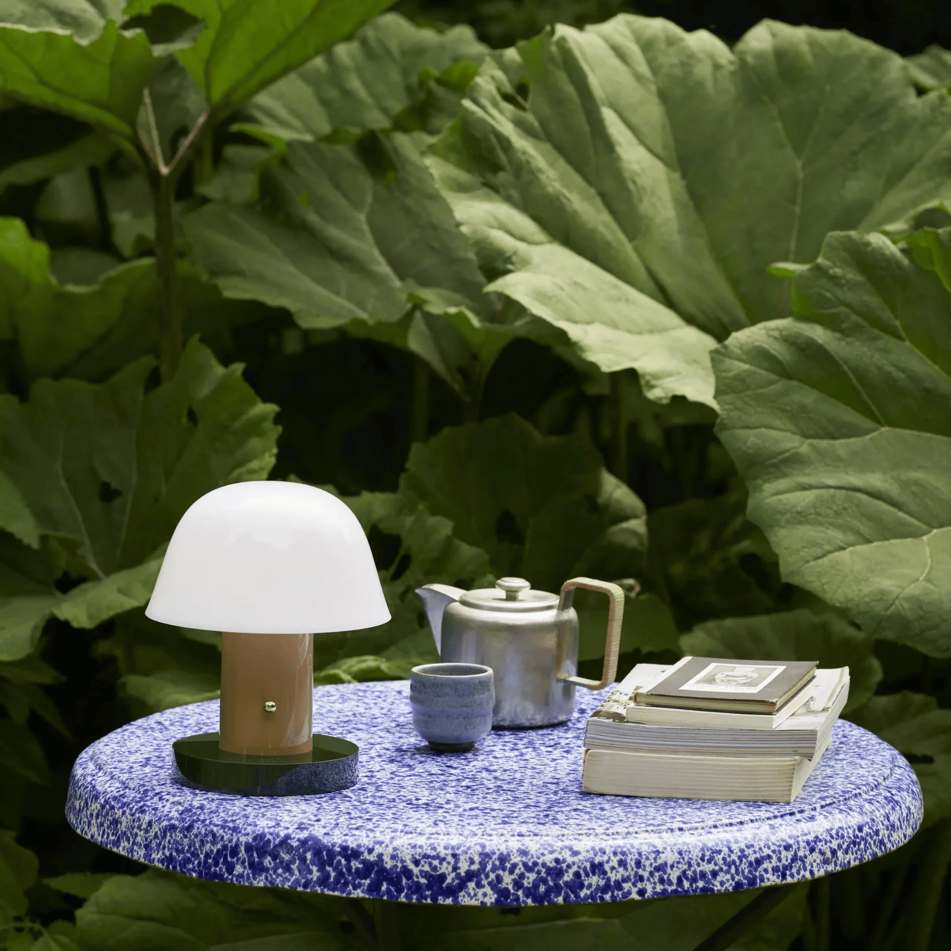 Table with a lamp, teapot, and books against a green leafy background
