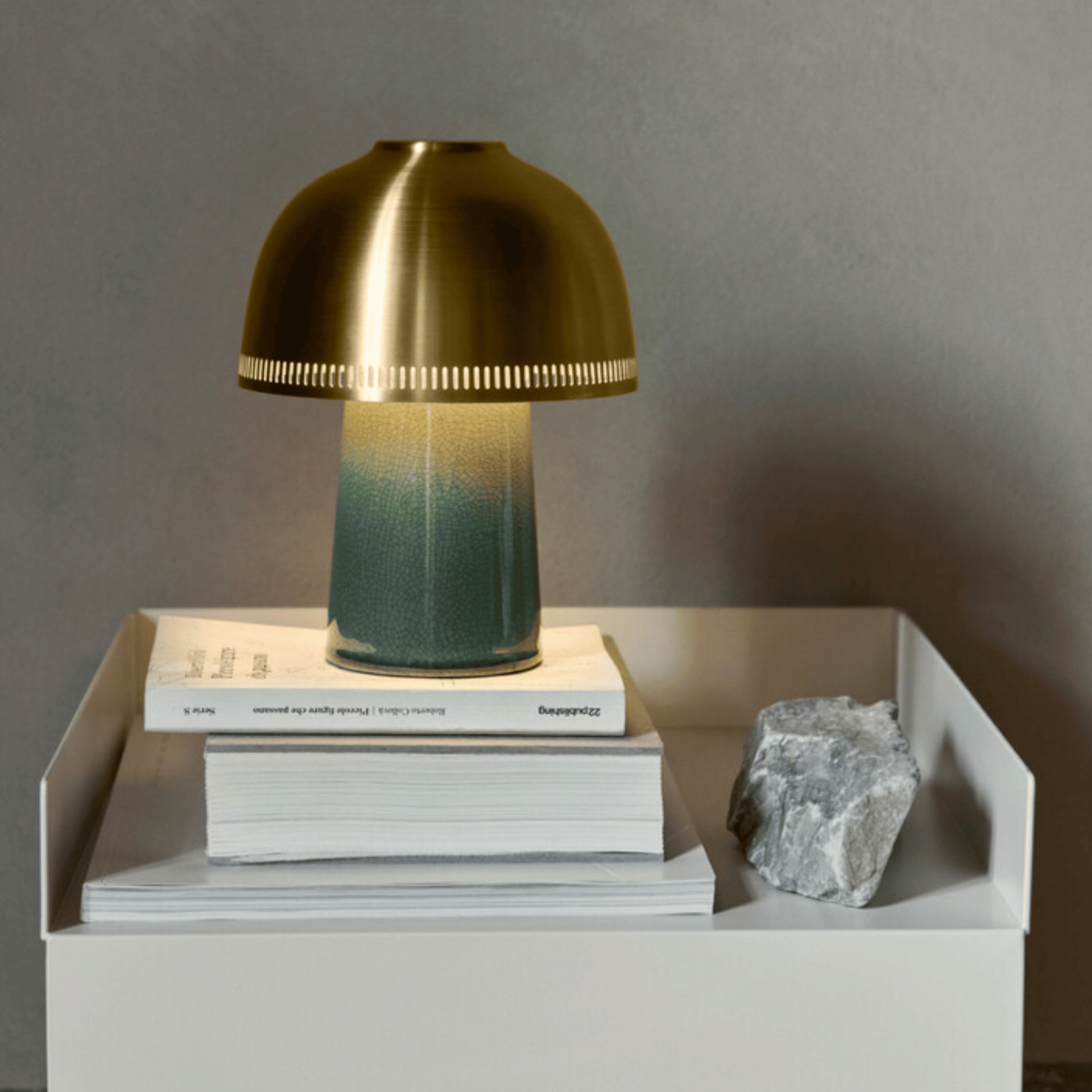 Brass and green gradient table lamp on a stack of books with a rock on a white surface.