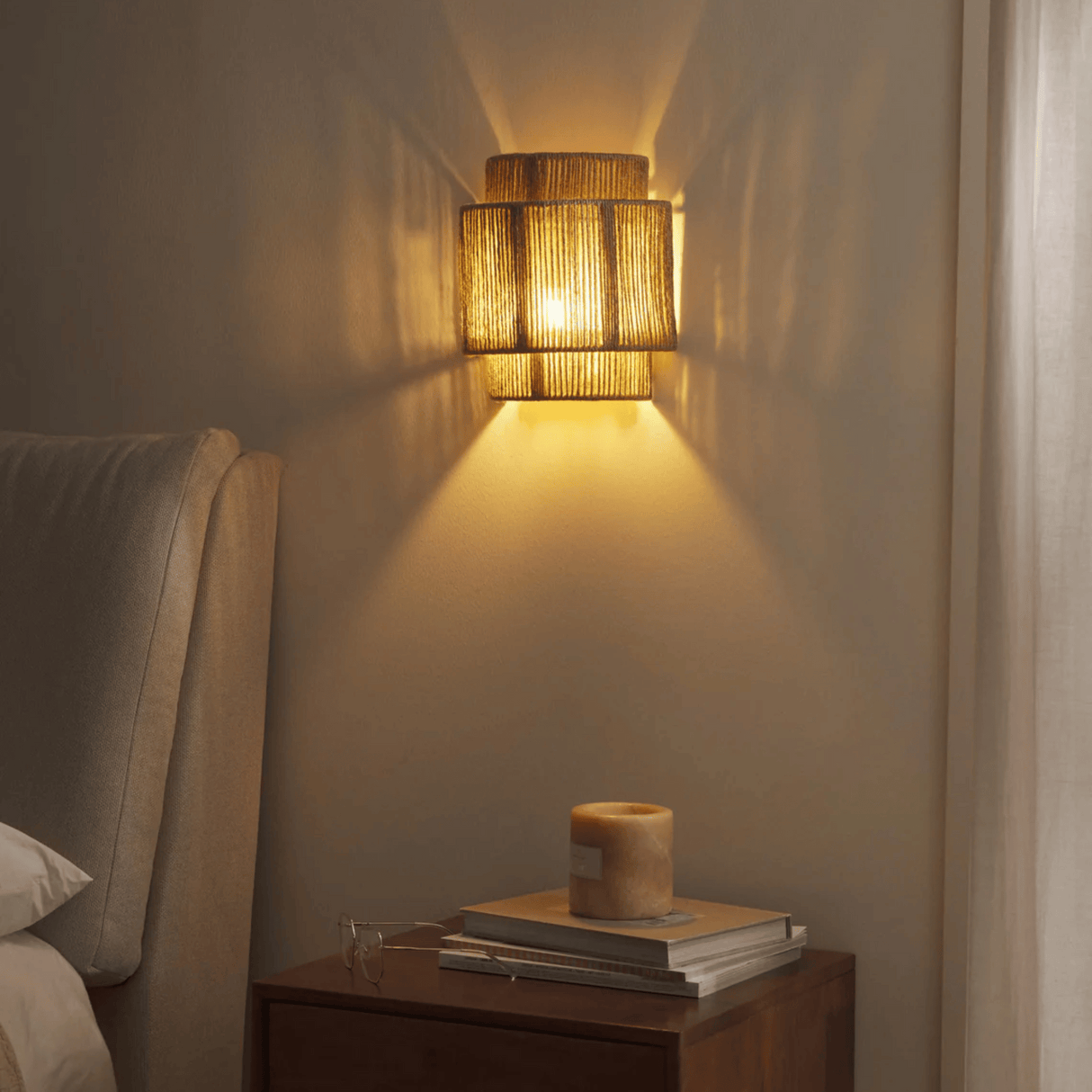 Woven wall light fixture casting a warm glow on a wall next to a nightstand with books and a candle.