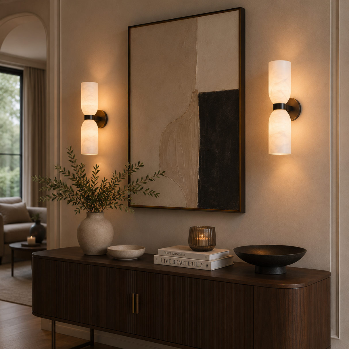 A pair of modern wall sconces with warm white cylindrical shades and dark metal bases are mounted on a beige wall on either side of a large abstract framed print. Below, a dark wood console table holds decorative vases, books, and bowls in a styled interior setting.