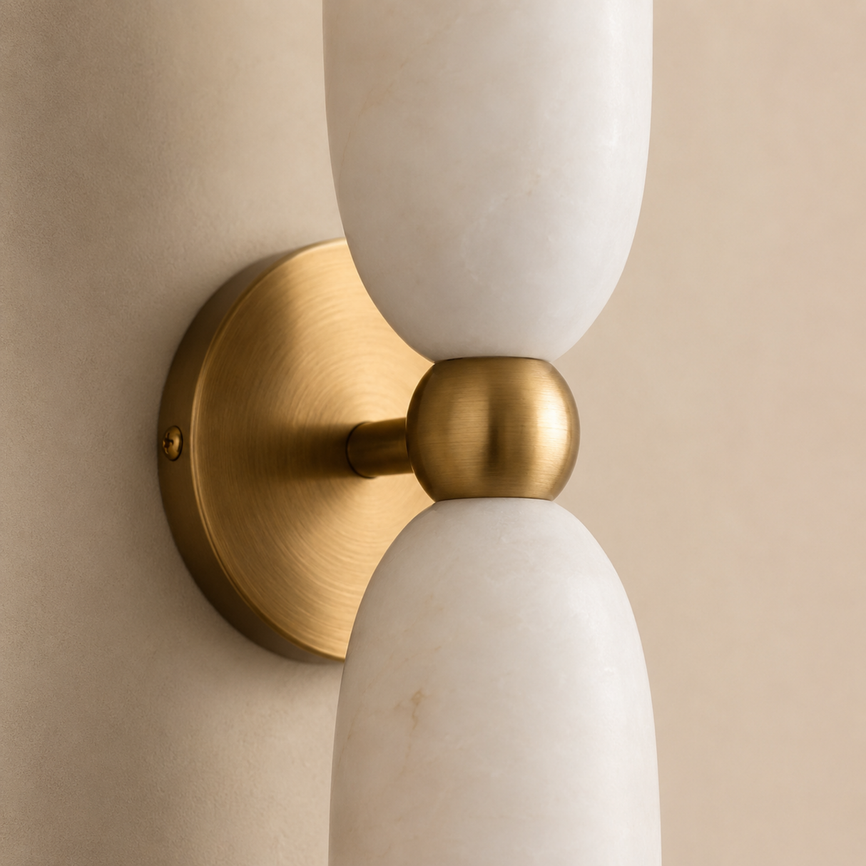 A modern wall-mounted light fixture with two elongated white marble shades and a brushed brass center joint and circular backplate, shown against a warm beige wall.