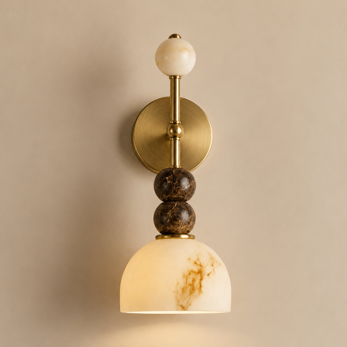A wall-mounted sconce with a round brass backplate, a vertical brass stem, a cream marble sphere at the top, two stacked dark brown marbled beads, and a dome-shaped cream marble shade with warm gold veining, shown against a beige wall.