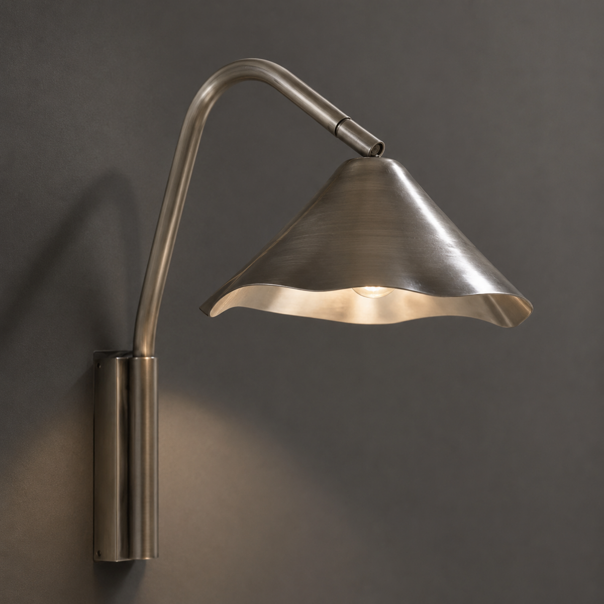 A modern wall-mounted sconce with a brushed metal finish, featuring a curved gooseneck arm and a flared shade that casts a warm light against a dark gray wall.