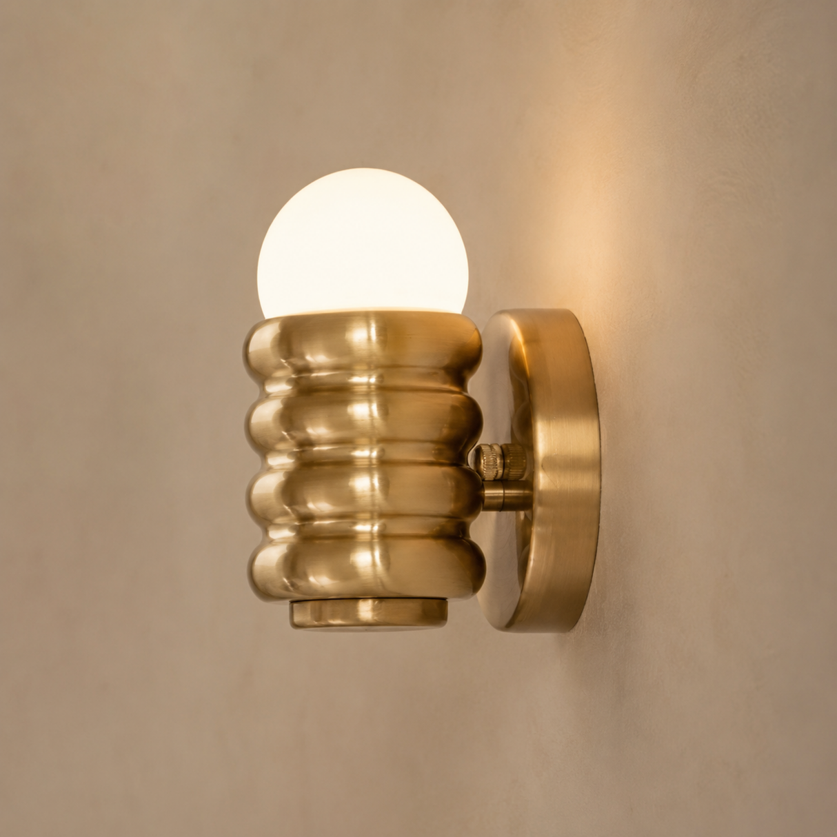 A modern wall-mounted sconce with a ribbed metallic gold shade and a glowing frosted globe bulb, mounted on a textured beige wall.