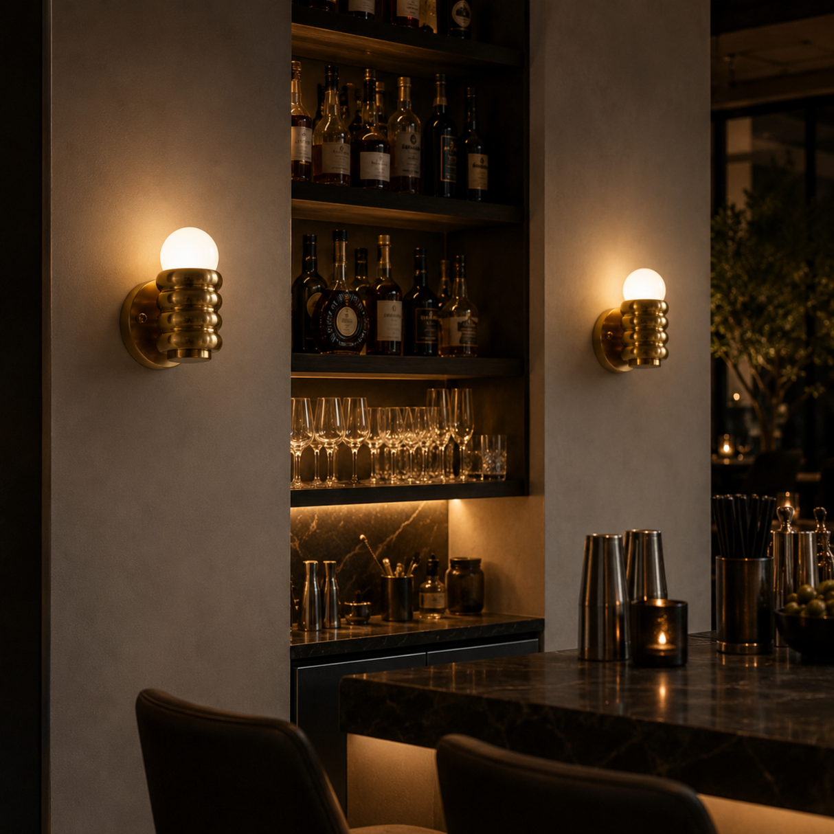 Two modern cylindrical wall sconces with textured gold-toned metal bases and glowing white globe bulbs, mounted on a warm beige wall in a dimly lit bar setting.