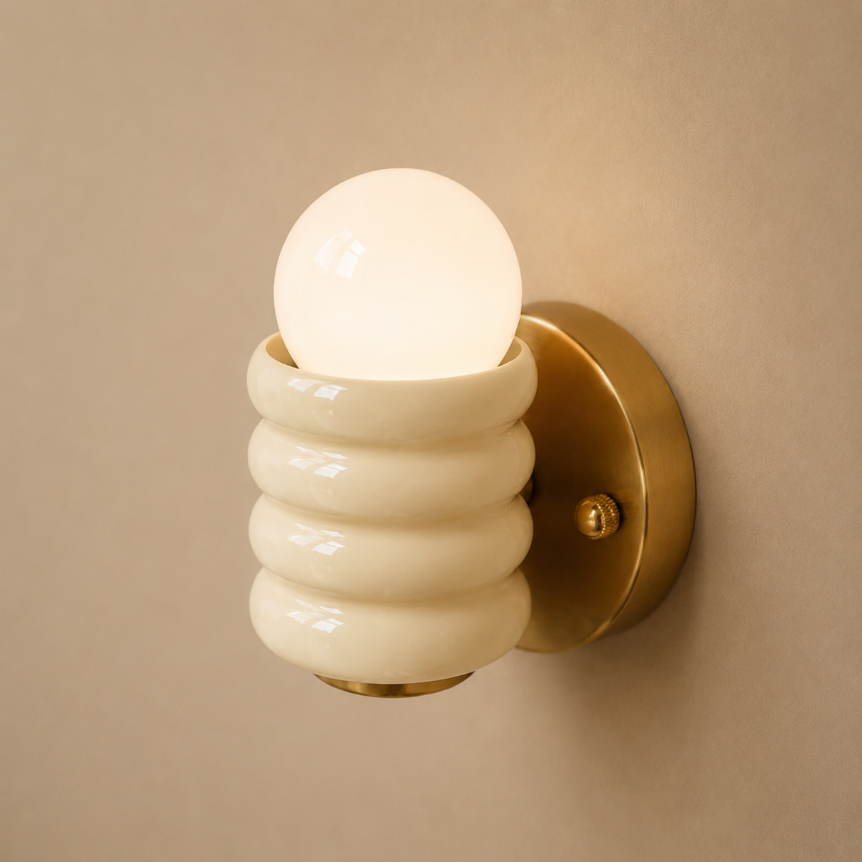 A modern wall-mounted sconce with a glowing frosted glass globe bulb, a ribbed cream-colored shade, and a brushed brass circular wall mount against a warm beige wall.