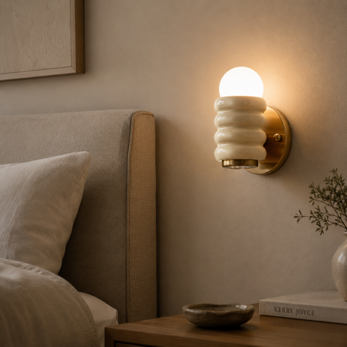 A warm wall-mounted sconce with a ribbed white shade and glowing frosted globe bulb, installed beside a bed in a softly lit bedroom.