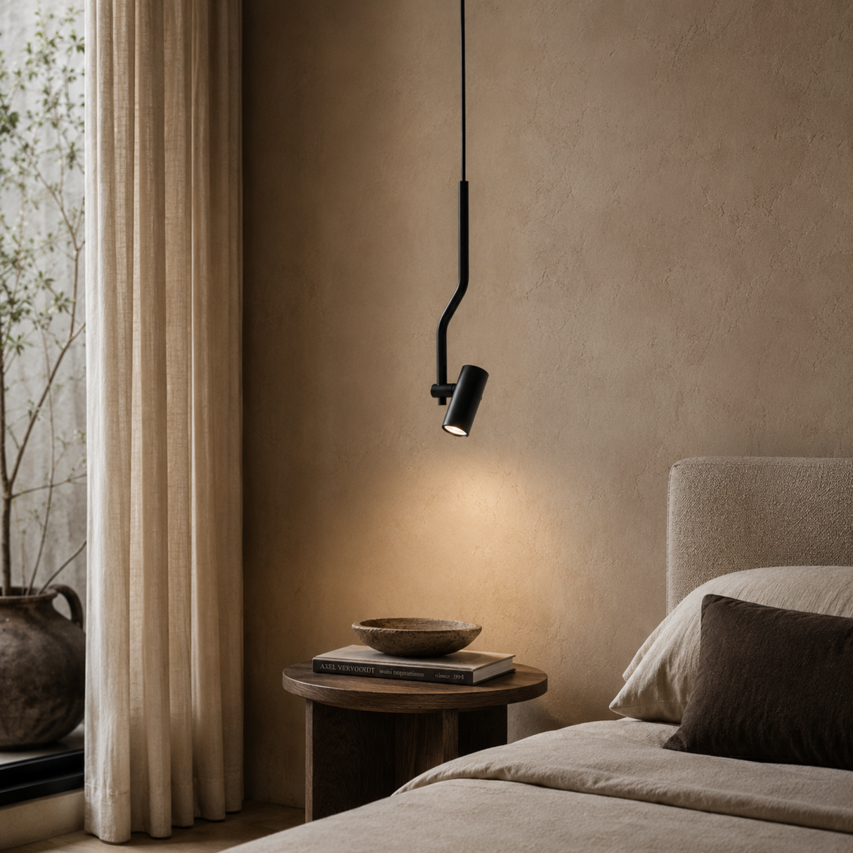 A modern black wall-mounted pendant light with a curved arm and adjustable cylindrical shade, casting a warm glow over a bed in a minimalist bedroom with beige walls, curtains, and a wooden side table.