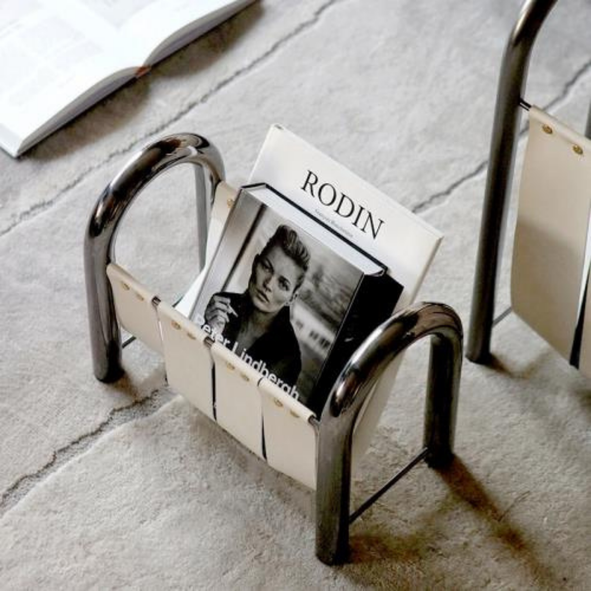 A white leather and chrome metal magazine holder on a gray carpet, holding a book with a black-and-white cover featuring a portrait.