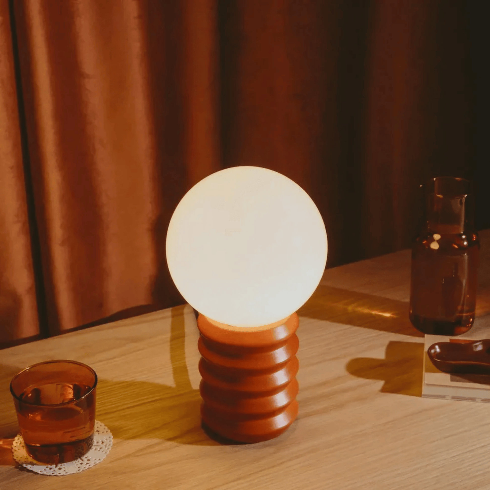 Table lamp with a textured brown base and white shade on a wooden table, with a glass of amber liquid and a book in the background.