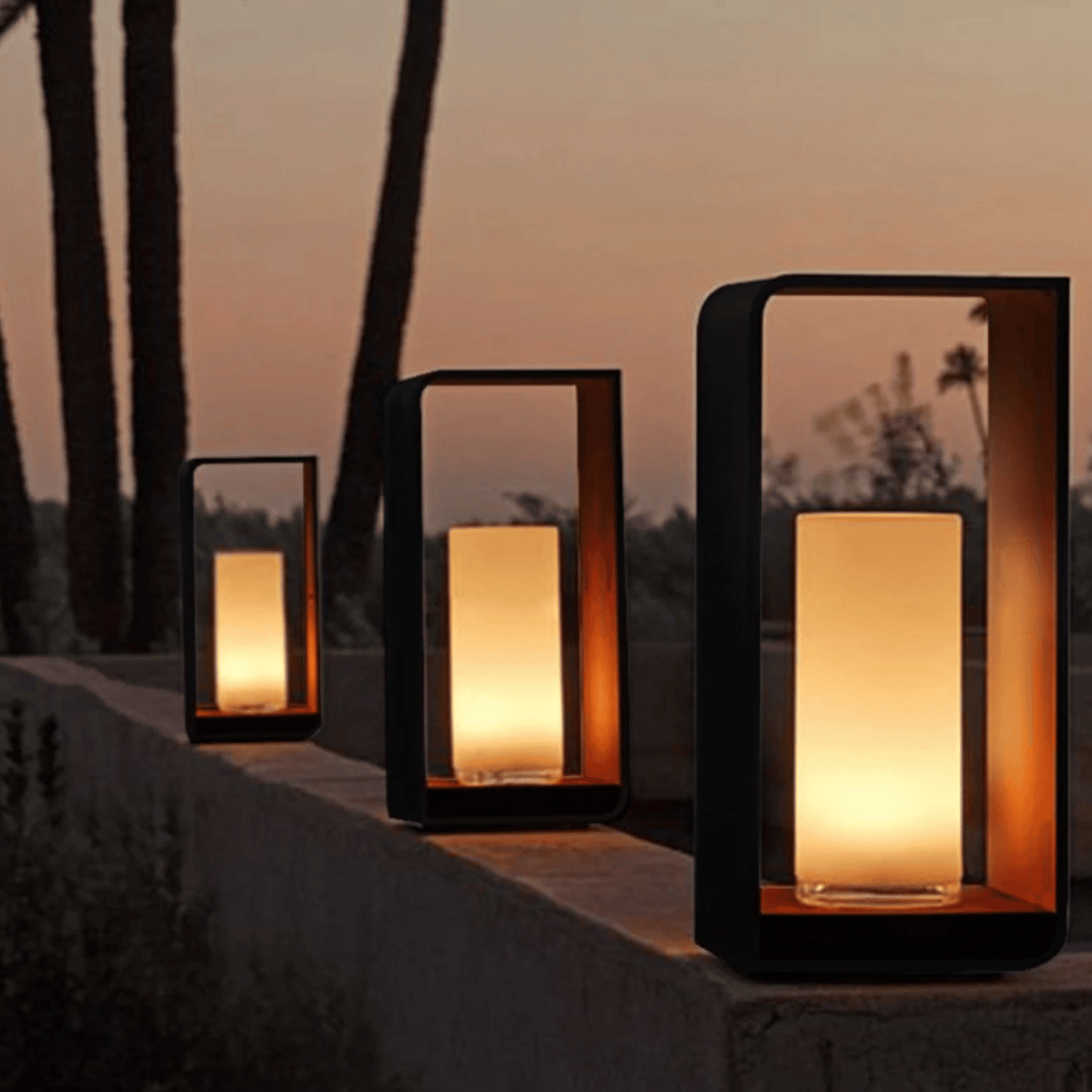Three rectangular outdoor lanterns with warm light on a stone ledge against a sunset sky.