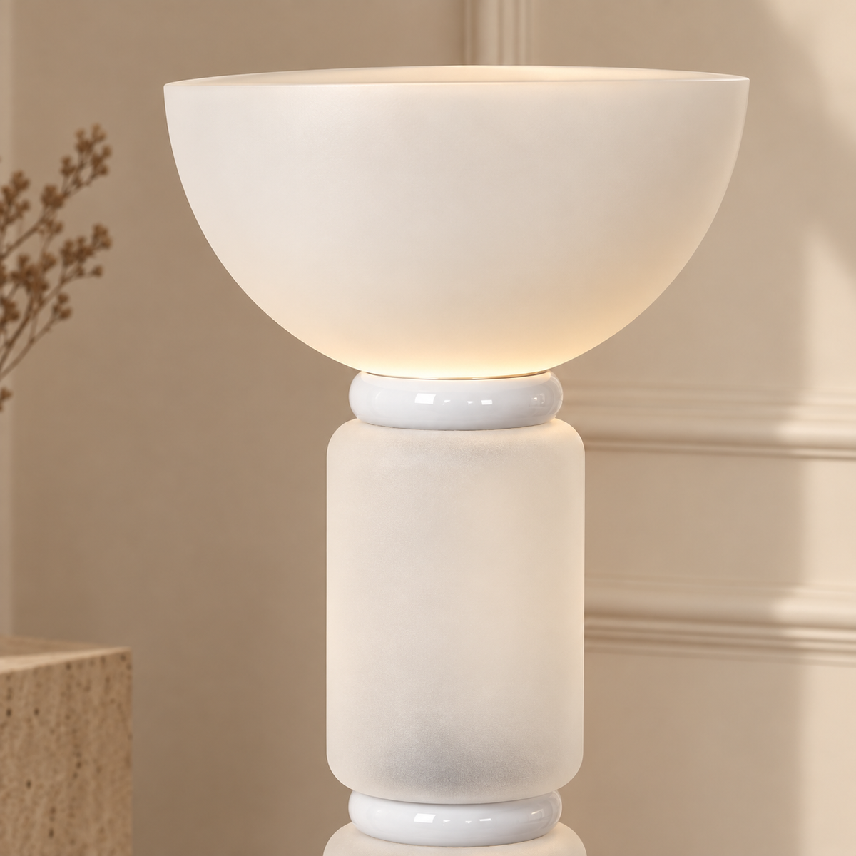 A modern table lamp with a wide, rounded bowl-shaped shade and a cylindrical base, both in a warm off-white or cream finish. The lamp is turned on, emitting a soft warm glow.