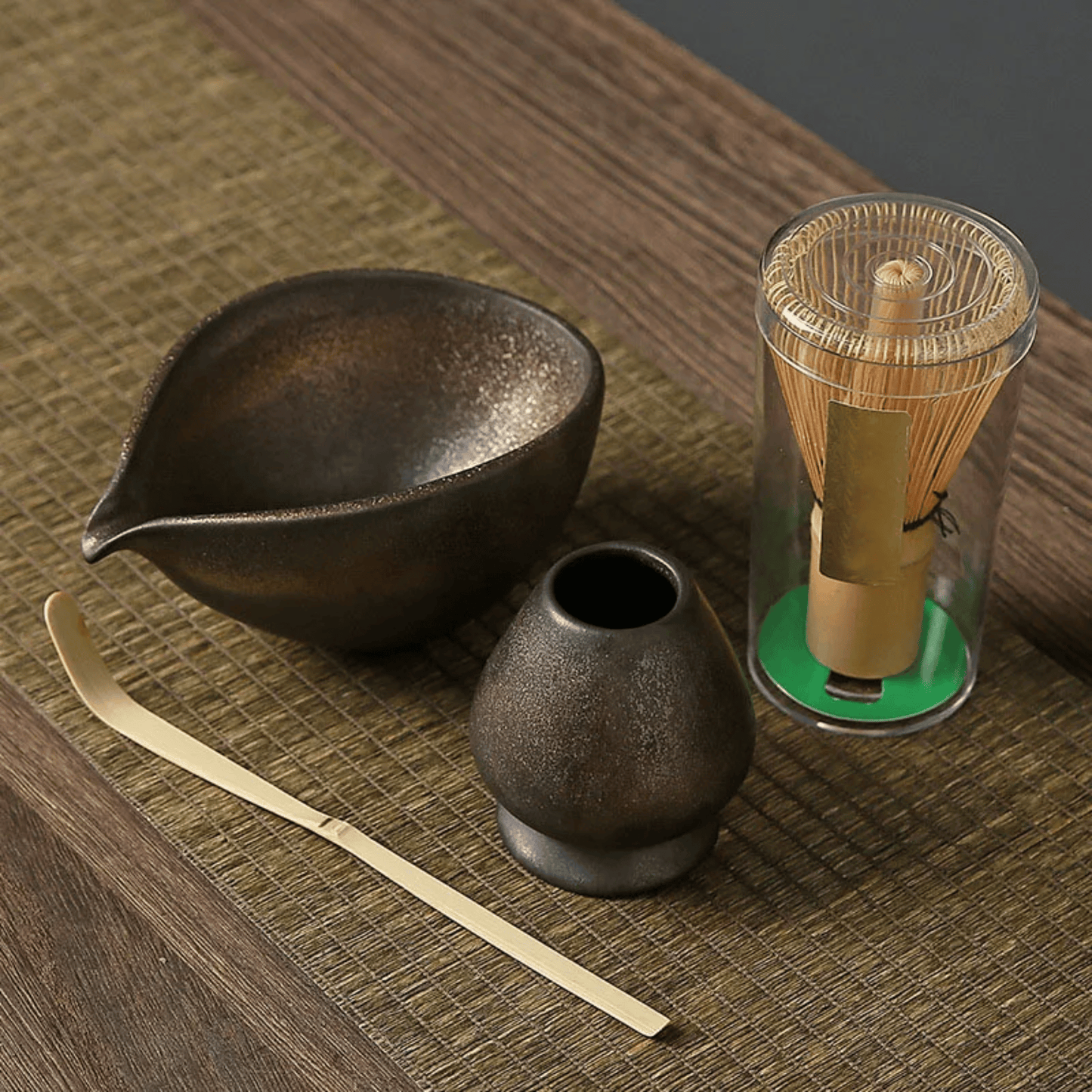 Tea-making set with black ceramic bowl, small container, gold whisk, and transparent container on a wooden surface.
