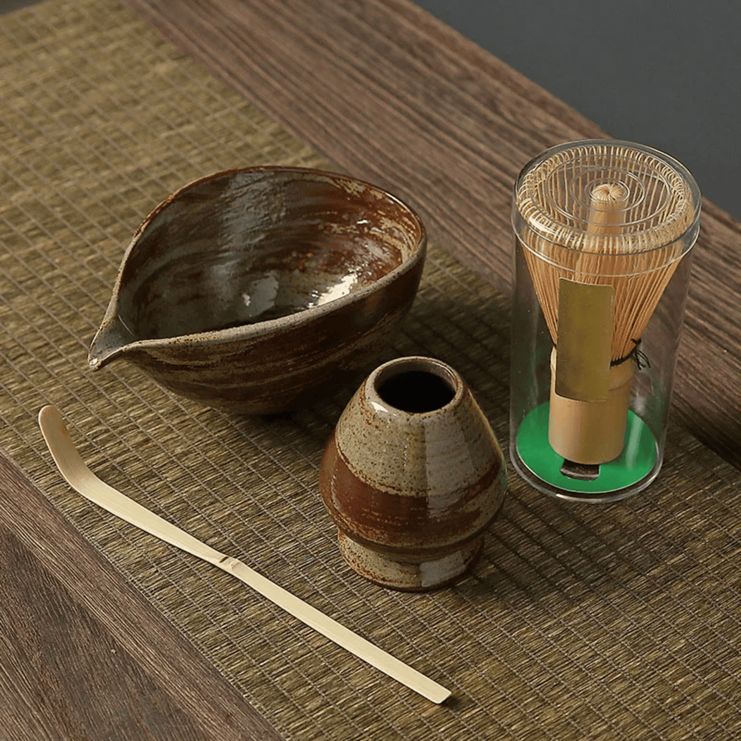 Tea-making set with ceramic bowl, whisk, scoop, and glass container on a wooden surface.