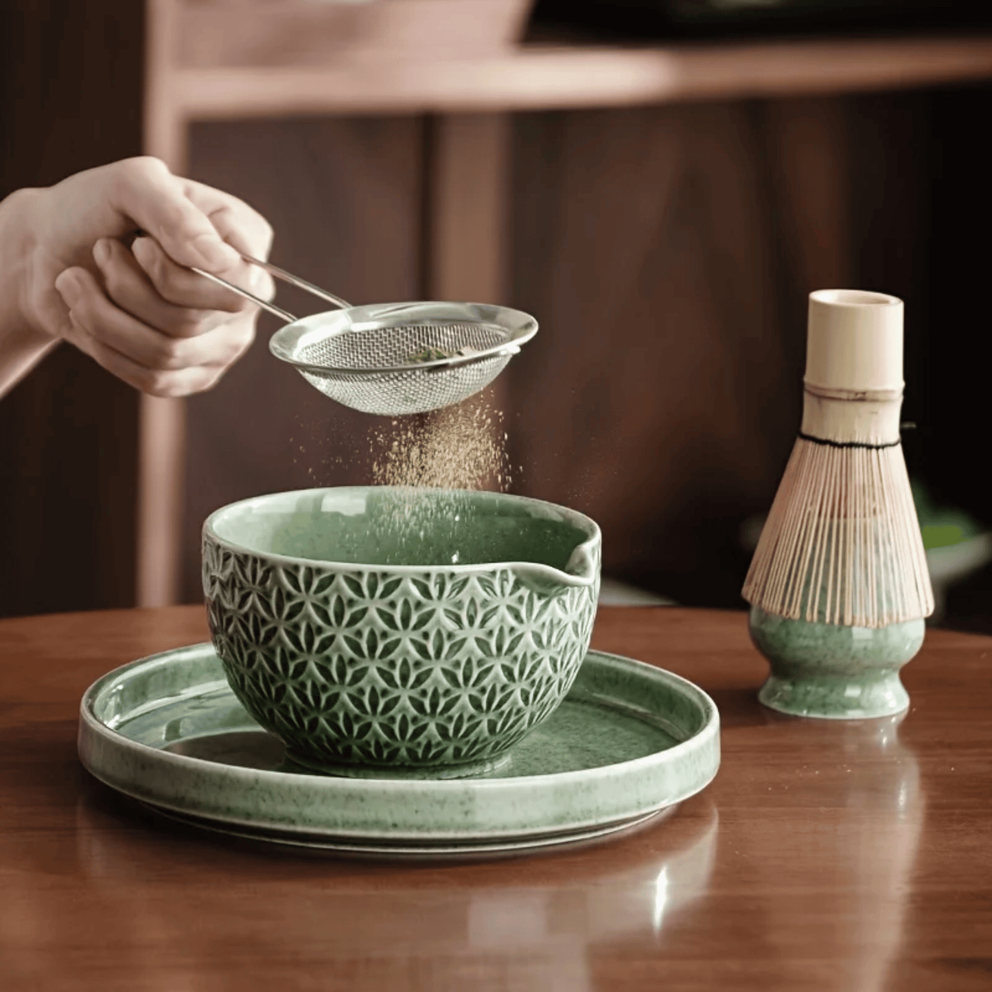 Person pouring tea leaves into a green ceramic bowl on a wooden table with a whisk in the background.