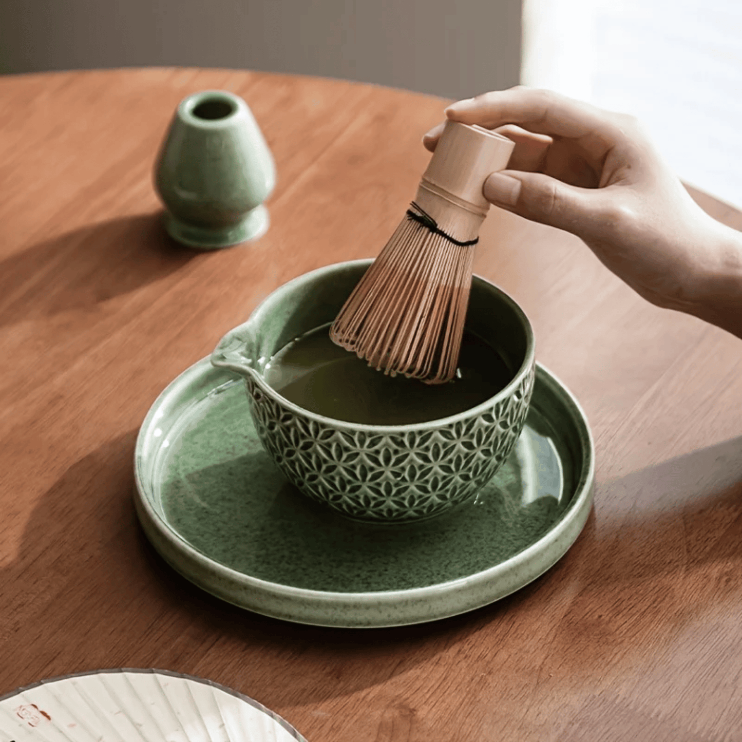 Person using a bamboo whisk to prepare matcha in a green ceramic bowl on a wooden table.