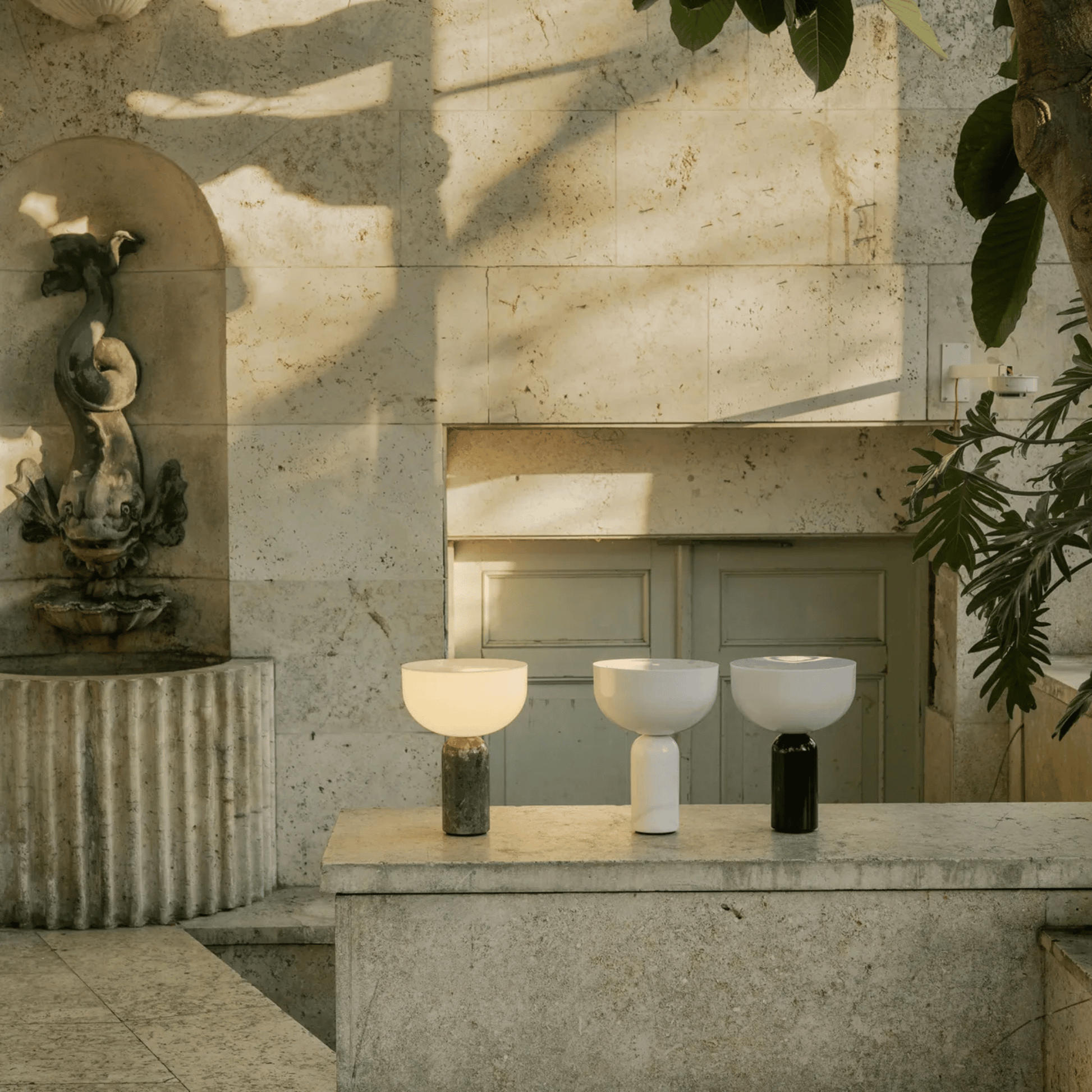 Three modern lamps on a stone surface with a fountain and stone wall in the background