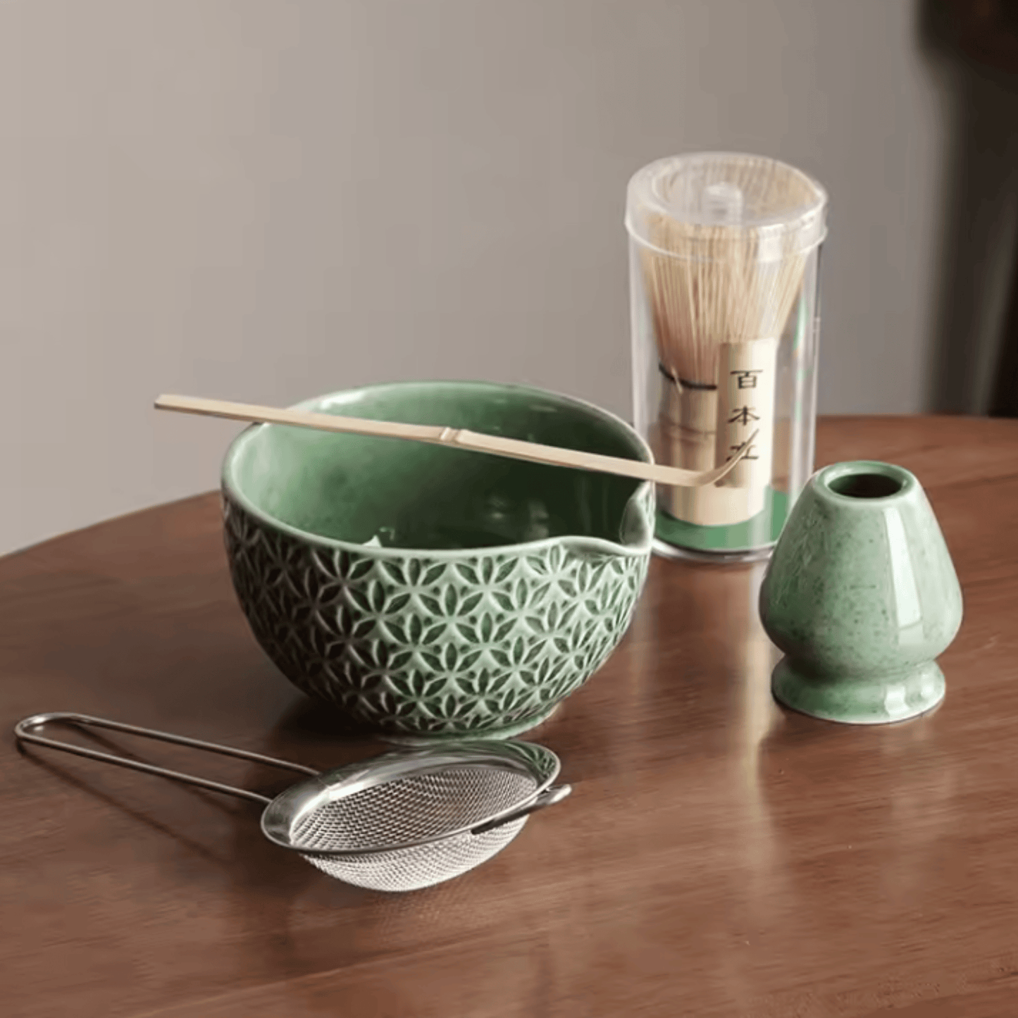Green ceramic bowl with pattern, wooden spoon, metal sieve, and glass container with bamboo sticks on a wooden surface.