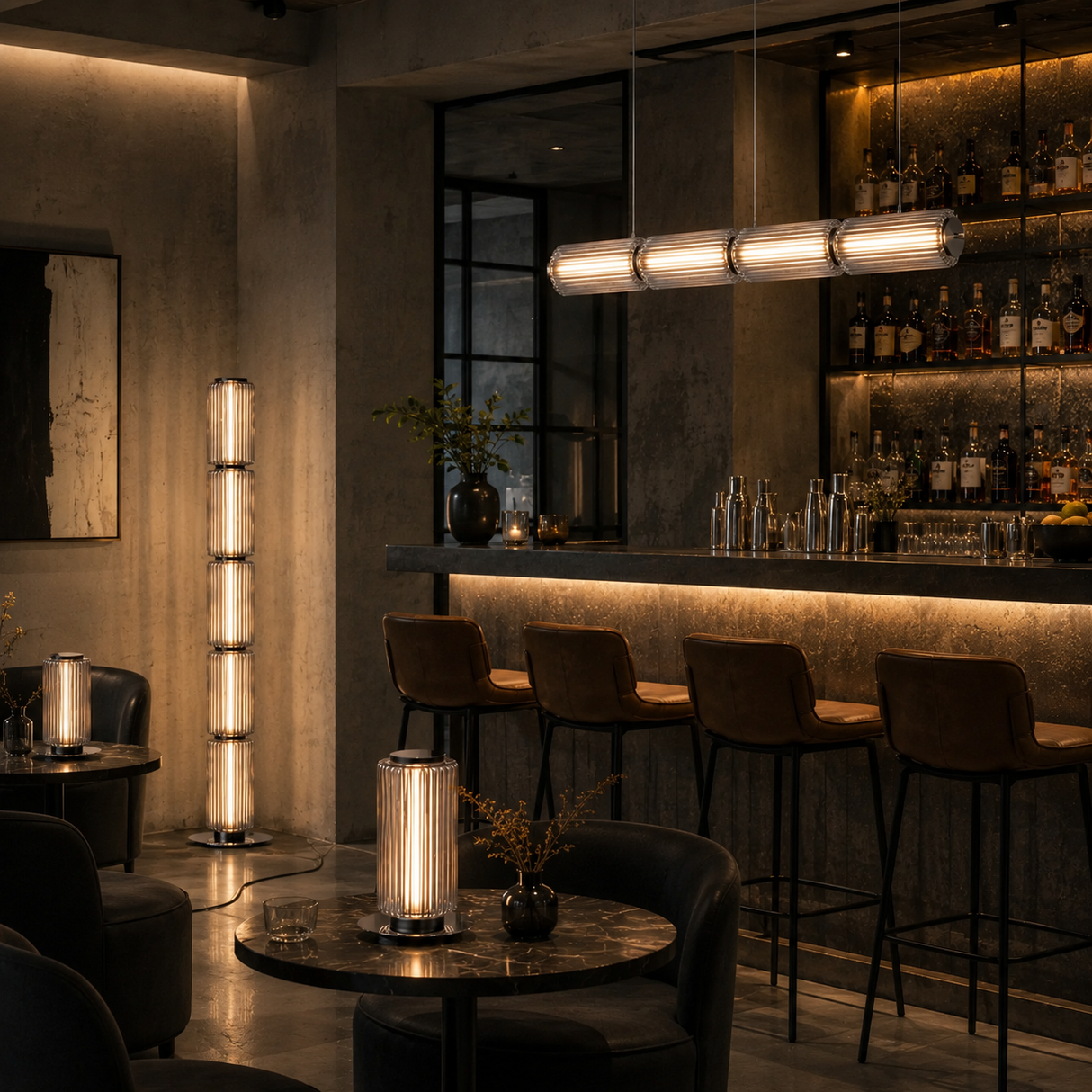 A modern bar interior with a tall cylindrical floor lamp, a matching table lamp on a round table, and a long horizontal pendant light over bar stools.