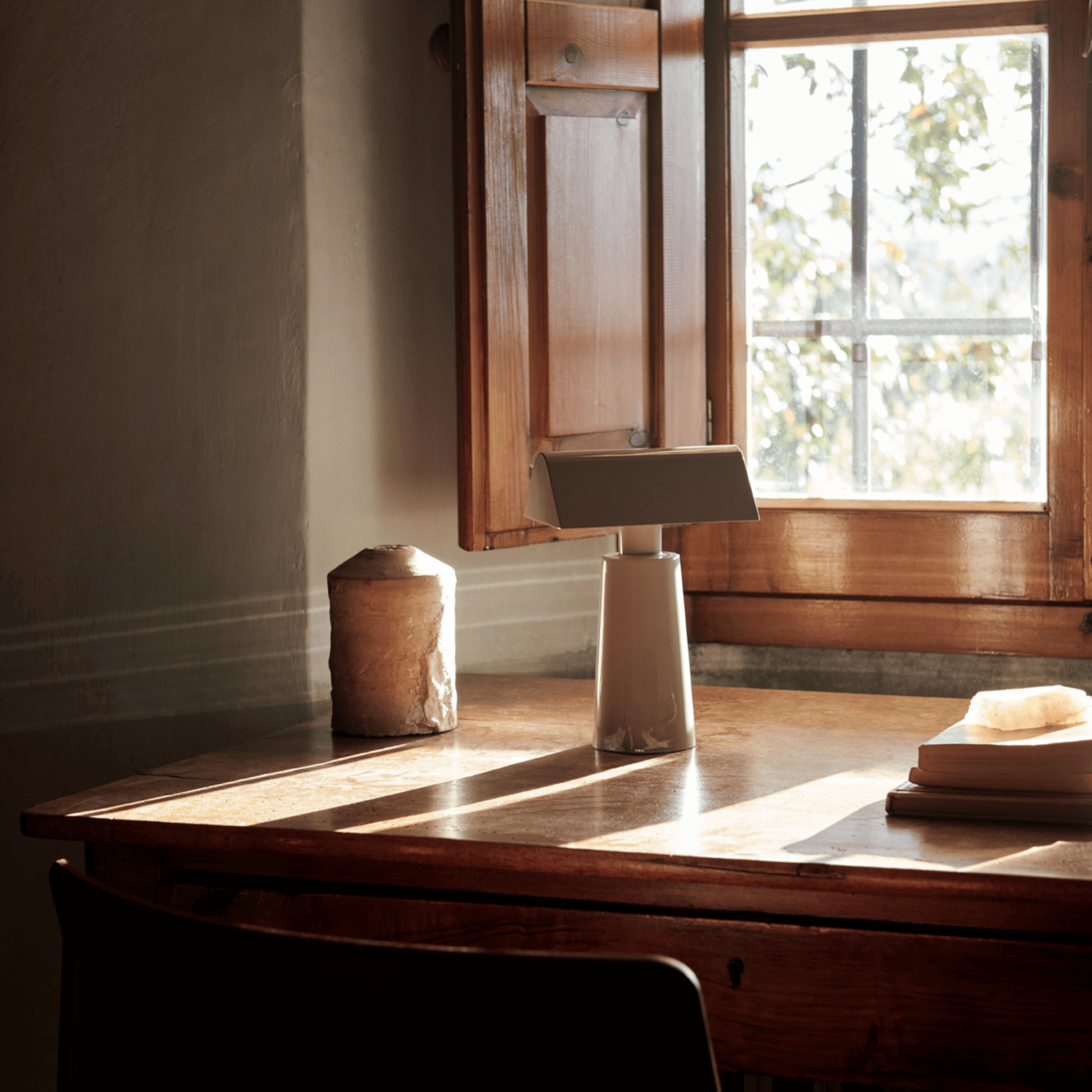 Wooden desk with lamp and objects near a window in a room with wooden shutters.