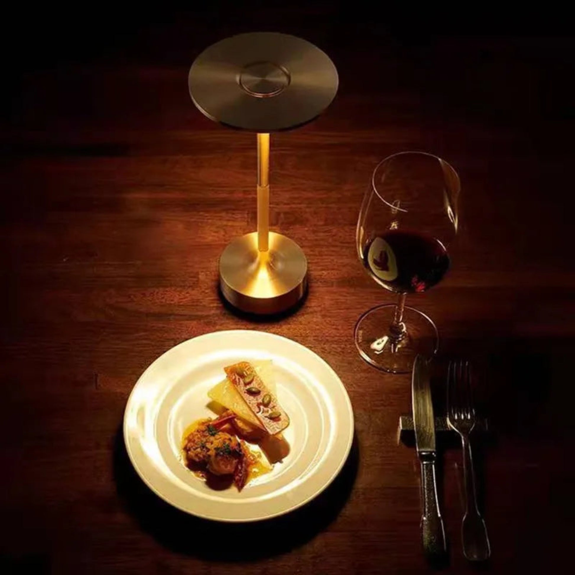 Dinner setting with a plate of food, wine glass, and table lamp on a wooden table.