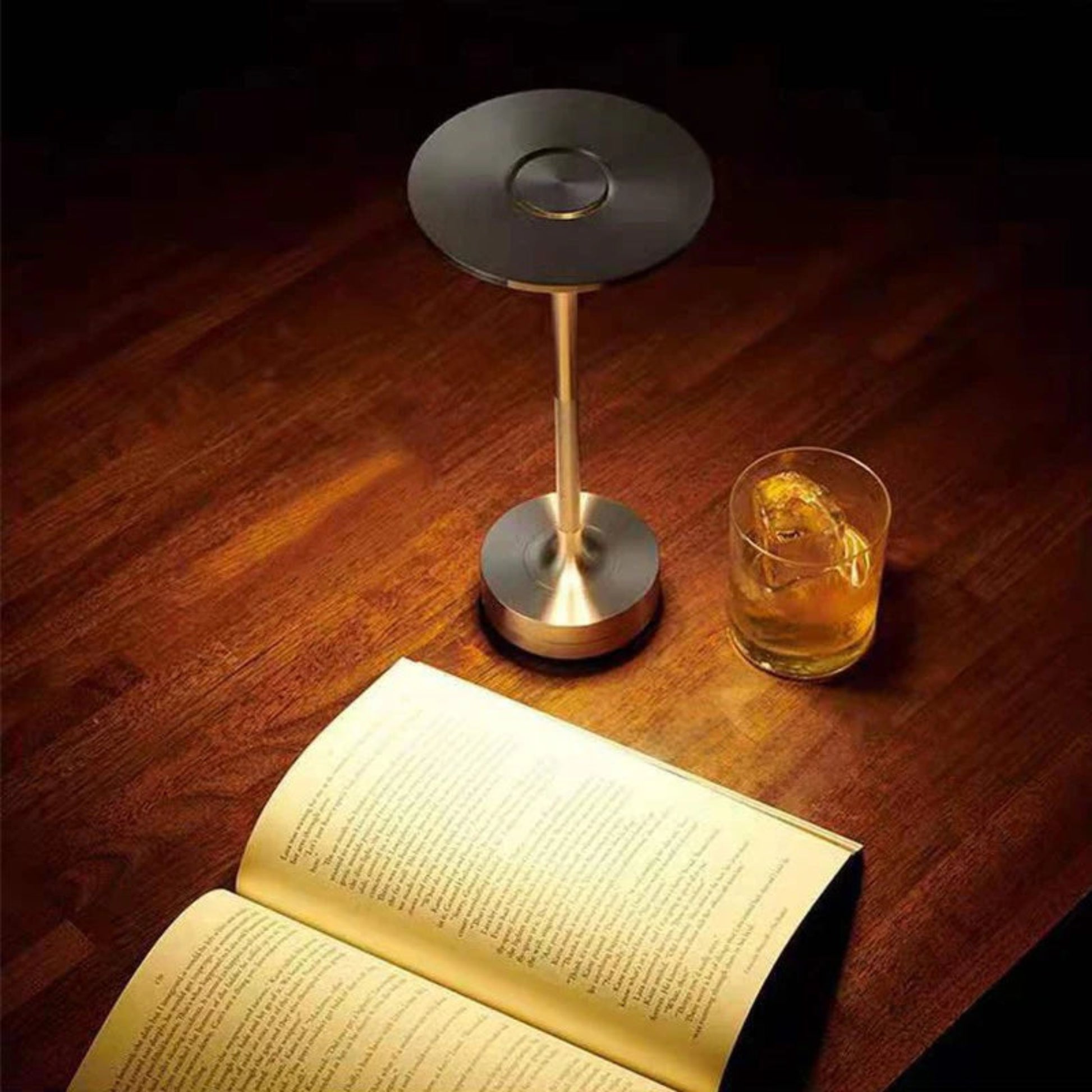 Small round table with a lamp, glass of whiskey, and open book on a wooden surface.