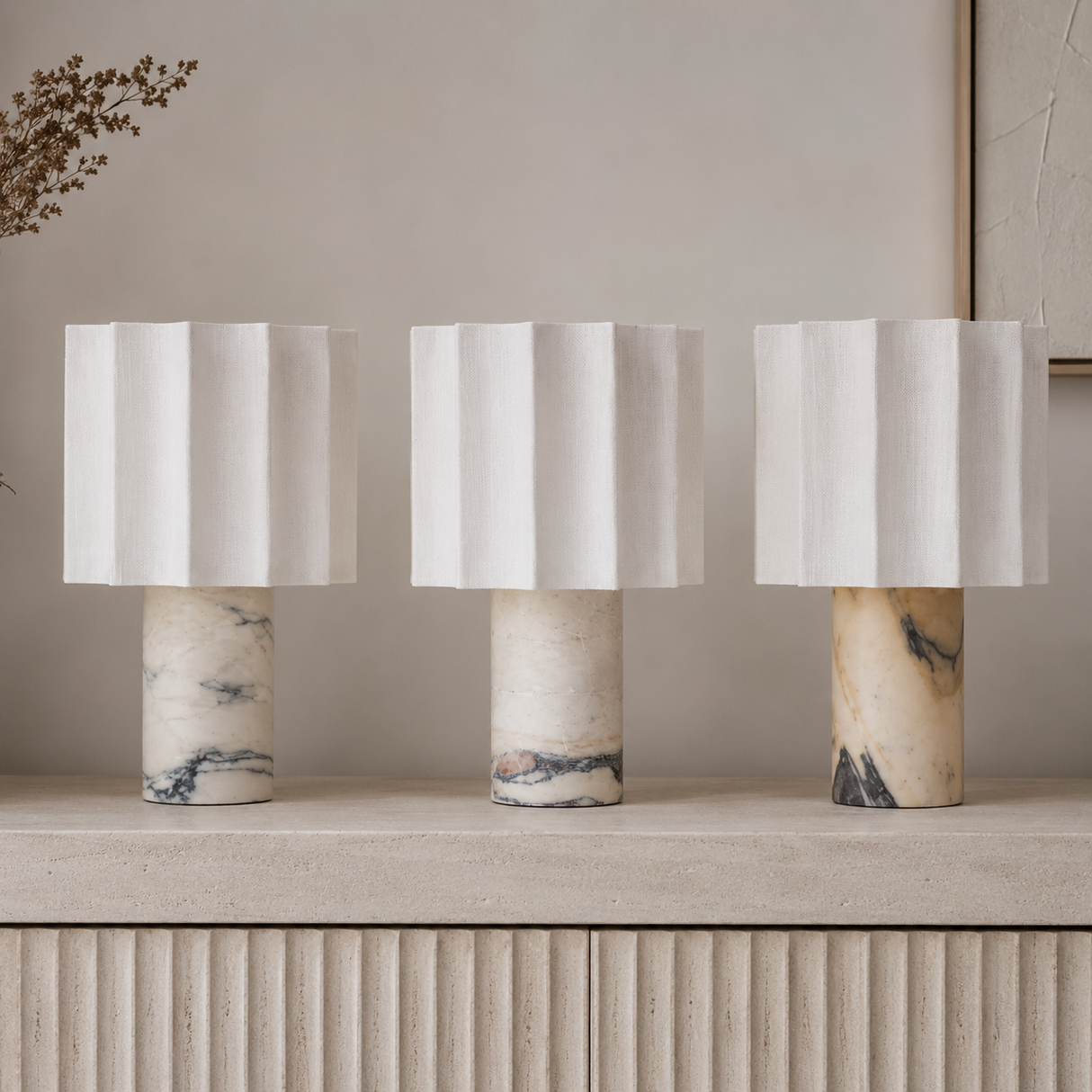 Three table lamps with white pleated drum shades and cylindrical marble bases in cream, beige, and gray tones, arranged on a light stone surface against a soft beige wall.