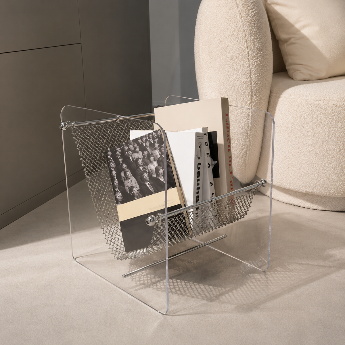 A clear acrylic floor magazine rack with a wire mesh front, holding several books and magazines, placed beside a cream boucle armchair in a modern interior setting.